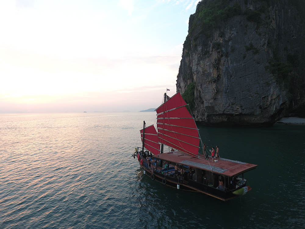 2 floor wooden tour boat in krabi sailing next to a cliff with its red sails raised at sunset