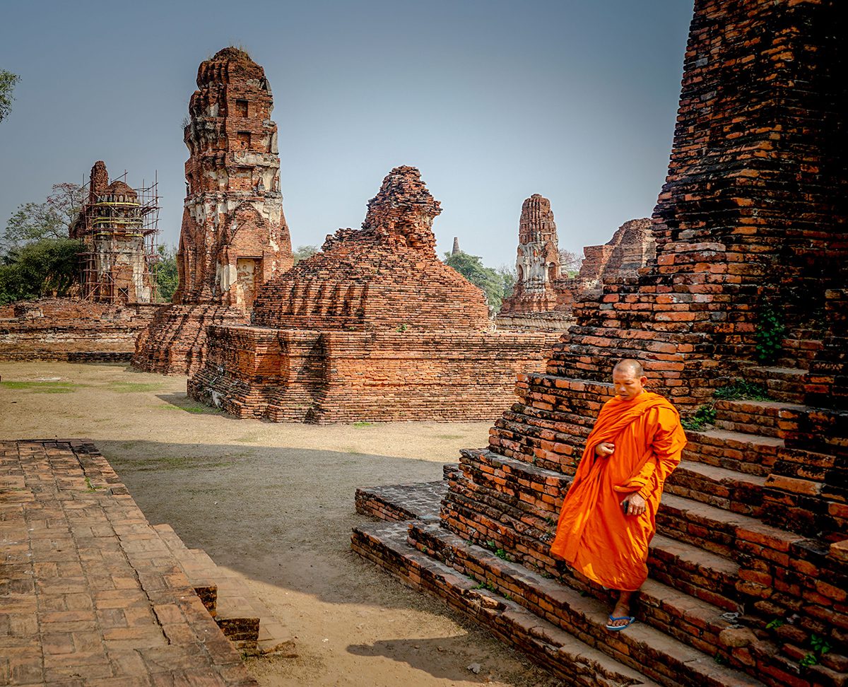 A monk wearing ornage robes walking down the steps of an old brick temple with other temples in the background