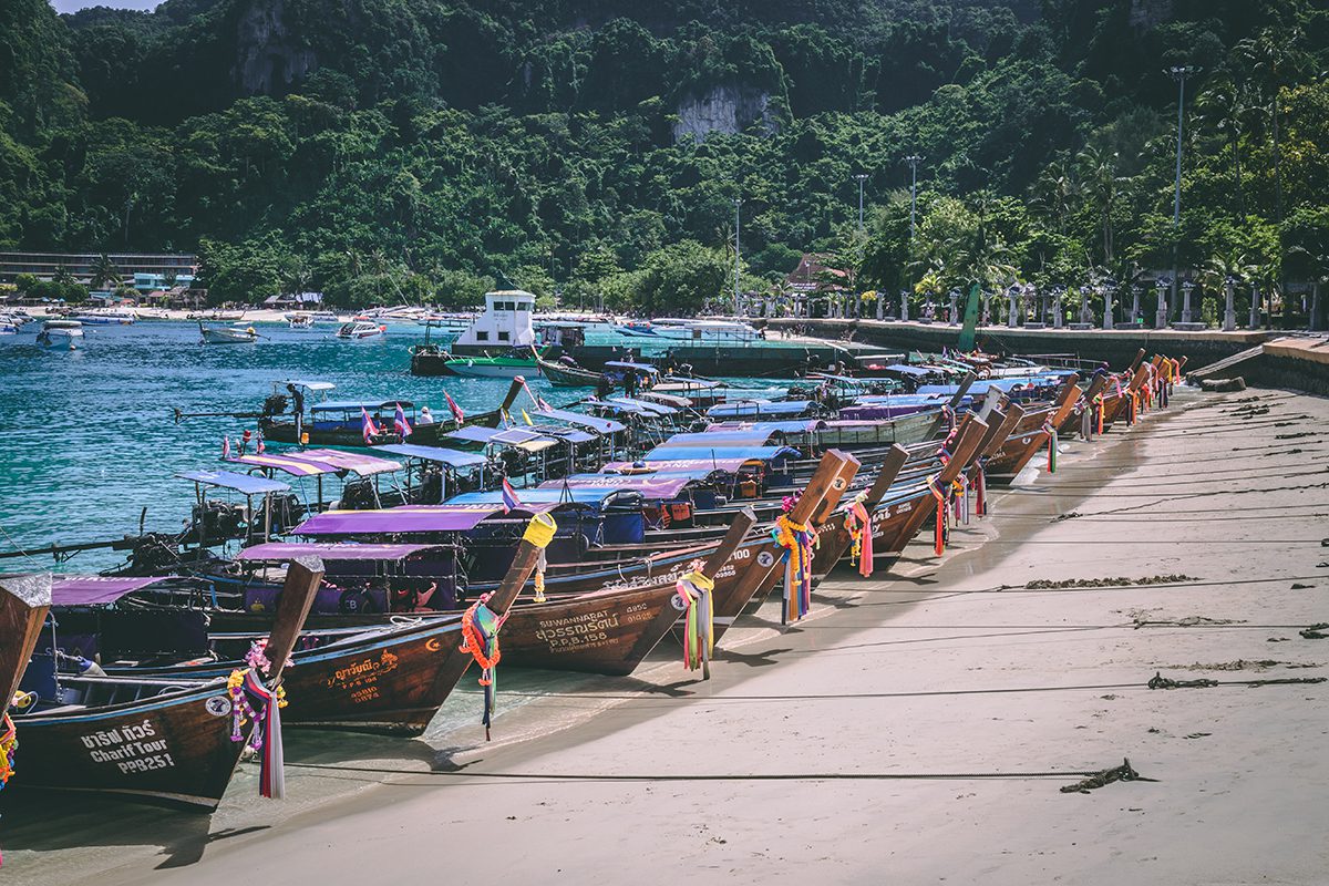 longtail boats in rows along a beach with the green tree-covered rocks in the background