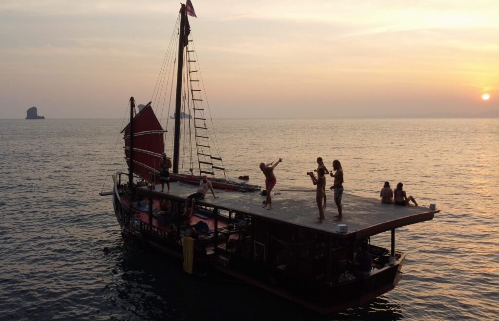a man about to jump from krabi sunset cruise pirate boat top deck while people watch and other watch the sunrise in the background
