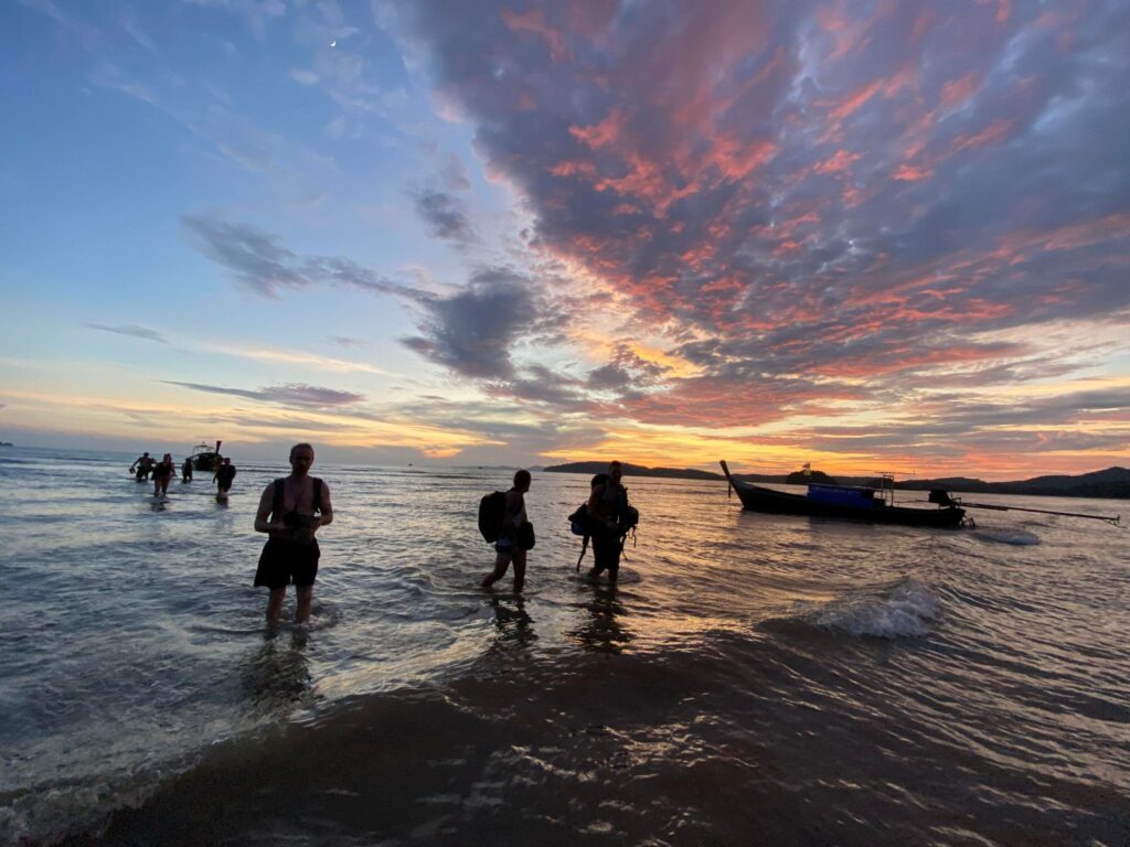 people wading through the water during sunset at aonang beach with colourful clouds of purple and orange
