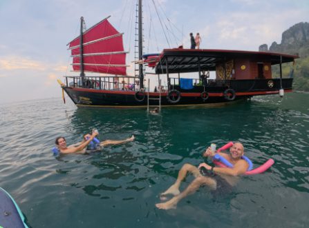 Guests floating near a red-sailed junk boat with limestone cliffs in the background