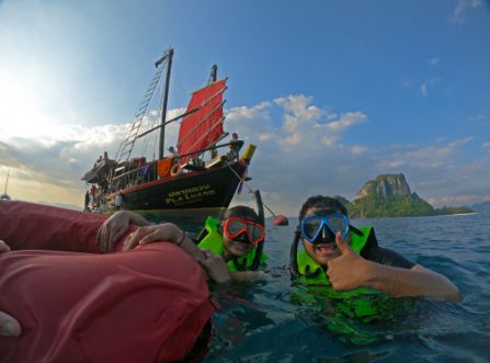 Two snorkelers in green life vests giving a thumbs-up near the red-sailed Pla Luang junk boat with limestone cliffs in the background