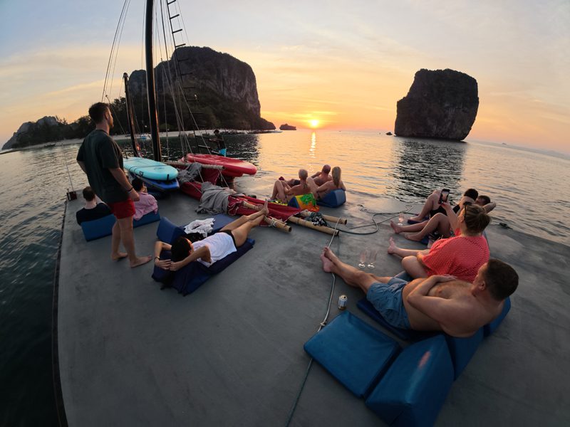 Group of guests lounging on cushions on the boat top deck, watching the sun set between Koh Poda and Koh Ta Ming.