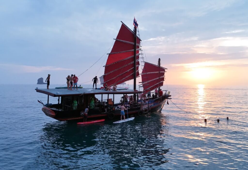 Krabi Sunset Cruises boat at sunset with guests paddleboarding next to it and others enjoying the sunset from the roof